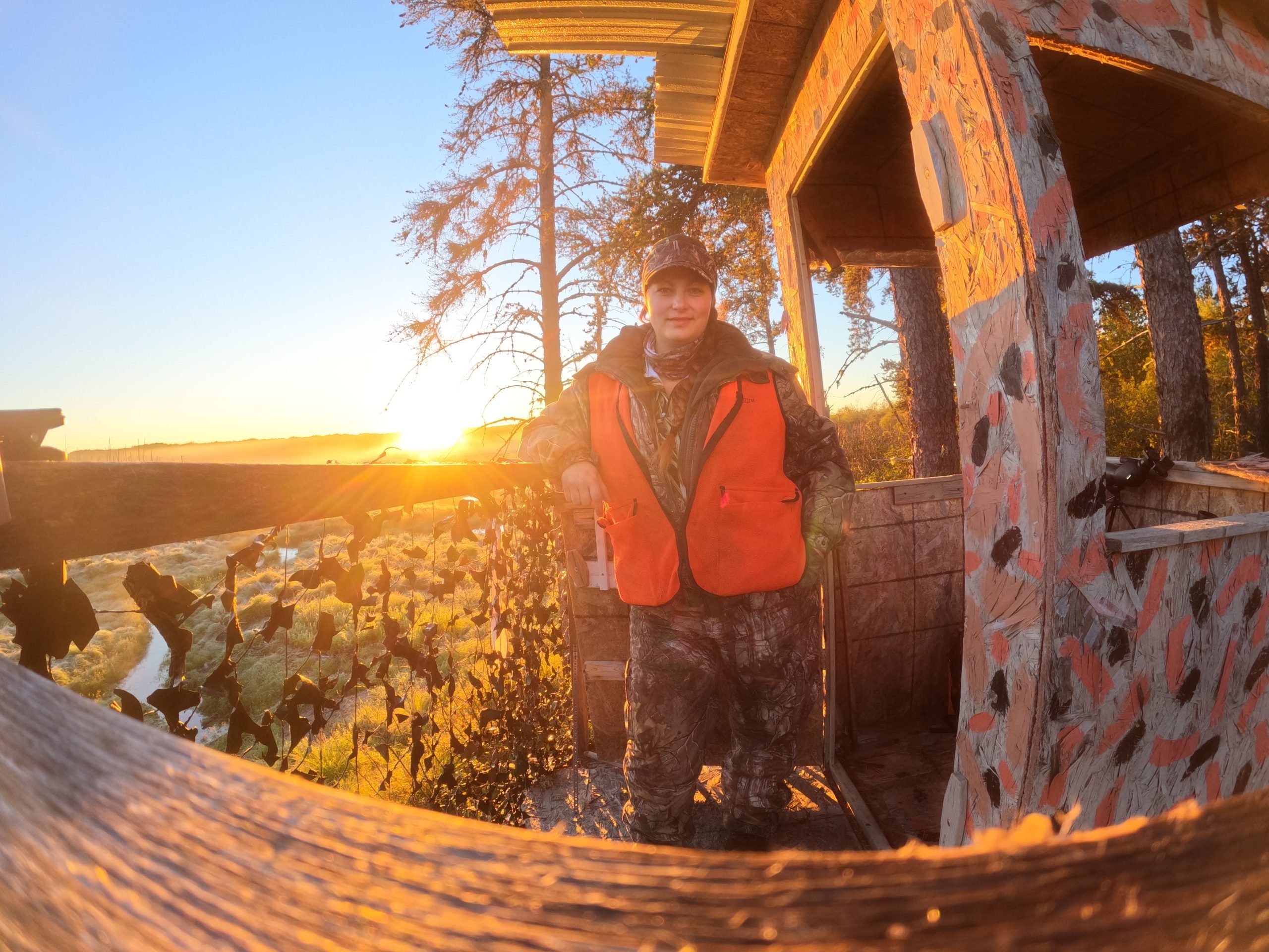 Stéphanie est debout dans sa tour de chasse, baignée dans une lumière jaune de fin de journée. Elle porte un habit de camouflage. 